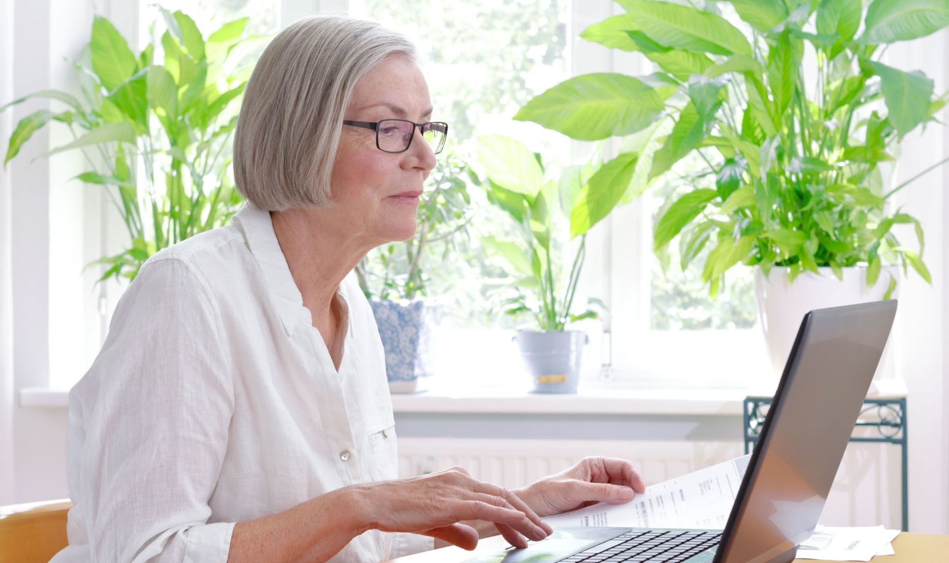 A woman wearing glasses uses a laptop beside a window